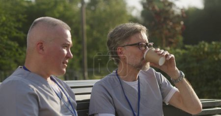 Photo for Two male doctors sit on the bench and drink coffee before start of work. Health care specialists discuss patient treatment outdoor. Mature physician helps colleague with making diagnosis. Close up. - Royalty Free Image