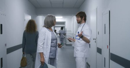 Téléchargez les photos : Spécialiste des soins de santé en uniforme blanc marche couloir du centre médical et parle avec une collègue féminine. Médecin au travail dans une clinique moderne. Personnel médical. - en image libre de droit