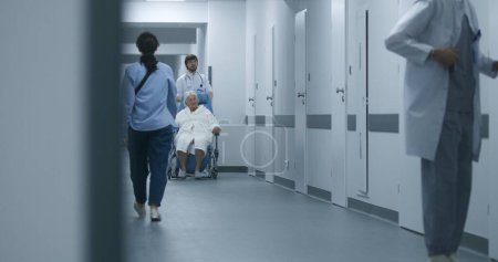 Foto de El doctor empuja una silla de ruedas con una paciente caminando por el pasillo de la clínica. Médico maduro abre la puerta, otro lleva a anciana a quirófano. Personal médico y pacientes en el pasillo del hospital. - Imagen libre de derechos