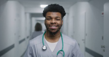 Photo for African American doctor stands in middle of medicine center hallway. Professional medic looks at camera and smiles. Caucasian doctor walks down the corridor with tests results. Medical staff at work. - Royalty Free Image