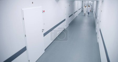 Photo for Bright clinic corridor: Female doctor and elderly woman walk down the hallway to hospital ward, talk about treatment. Medical staff work in modern medical facility. Security camera view. High angle. - Royalty Free Image