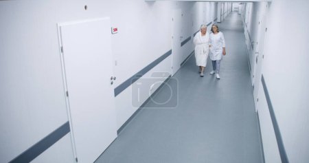 Téléchargez les photos : Couloir lumineux de clinique : Femme médecin et femme âgée marchent dans le couloir à la salle d'hôpital, parlent du traitement. Le personnel médical travaille dans un établissement médical moderne. Vue caméra de sécurité. Angle élevé. - en image libre de droit