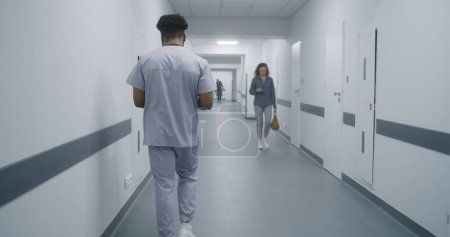 Photo for African American health care specialist walks hospital hallway. Male doctor goes on appointment with patients. Multi ethnic medical personnel at work in modern clinic. - Royalty Free Image