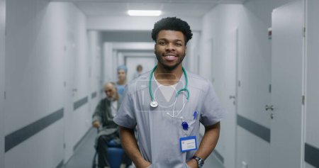 Photo for African American doctor looks at camera and smiles. Young doctor stands in middle of hospital corridor. Nurse transports senior patient in wheelchair in medical room. Medical staff at work in clinic. - Royalty Free Image