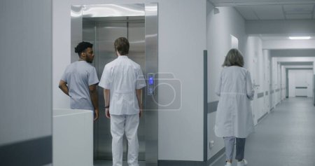 Téléchargez les photos : Médecin afro-américain vient lever la porte avec un collègue et appuie sur le bouton d'appel. Les travailleurs médicaux attendent ascenseur dans le couloir du centre médical. Spécialistes multiraciaux de la santé au travail à l'hôpital. - en image libre de droit