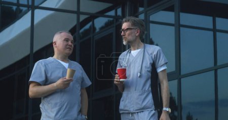 Photo for Two doctors in uniform stand near medical center entrance in the evening. Professional medics drink coffee, discuss work. Medical staff of hospital or modern clinic. Lifestyle and healthcare concept. - Royalty Free Image
