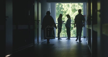 Photo for Hospital hallway: Doctors and professional medics walk. Nurse with digital tablet stands with adult woman near window and talks. Medical staff and patients in dark clinic or medical center corridor. - Royalty Free Image