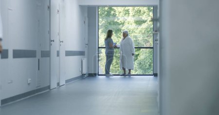 Photo for Hospital hallway: Doctors and professional medics walk. Nurse with digital tablet comes to elderly female patient standing near window. Medical staff and patients in clinic or medical center corridor. - Royalty Free Image