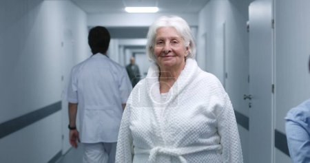 Foto de Mujer mayor está en el corredor de la clínica moderna. Paciente anciana sonríe y mira a la cámara. Personal médico, médicos y pacientes en el fondo en el pasillo del hospital o centro médico. Retrato. - Imagen libre de derechos