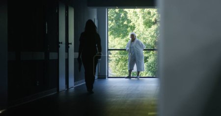Photo for Clinic corridor: Doctors and professional medics walk. Nurse with papers comes to elderly female patient standing near window. Medical staff and patients in dark hospital or medical center hallway. - Royalty Free Image