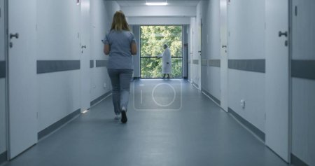 Photo for Clinic corridor: Doctors and professional medics walk. Nurse with digital tablet comes to elderly woman standing near window. Medical staff and patients in modern hospital or medical center hallway. - Royalty Free Image