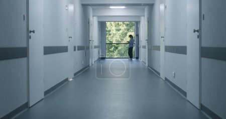 Photo for Clinic corridor: Doctors and professional medics walk. Nurse with digital tablet comes to female patient standing near window. Medical staff and patients in modern hospital or medical center hallway. - Royalty Free Image