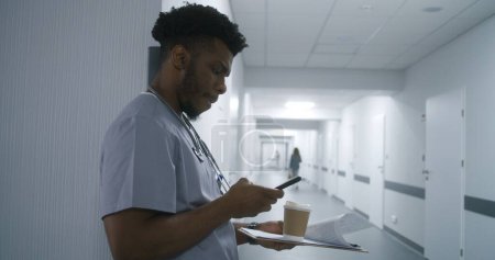 Photo for African American healthcare specialist with coffee and folder stands in hospital corridor leaning wall and takes phone. Doctor waits appointment with patient in clinic hallway. Medical staff at work. - Royalty Free Image