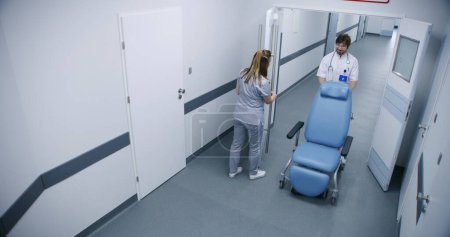 Photo for Clinic corridor: Doctors, professional medics and patients walking. Male nurse pushes wheelchair. Elderly man goes to his hospital room. Medical staff work in modern medical facility. High angle. - Royalty Free Image
