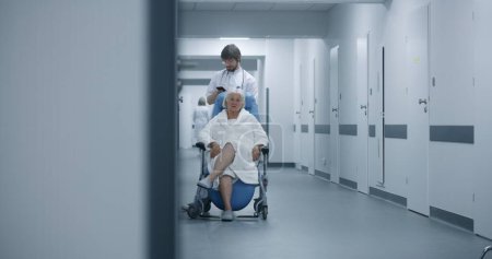 Photo for Male doctor stands with elderly woman in wheelchair in clinic corridor, uses mobile phone. Medic with female patient near operating or procedures room. Medical staff and patients in hospital hallway. - Royalty Free Image