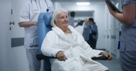 Photo for Elderly woman in wheelchair talks to doctor in clinic corridor. Medic discusses tests results with patient, uses digital tablet. Medical staff and patients walk in background in hospital hallway. - Royalty Free Image