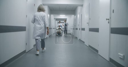 Photo for Professional doctors transport elderly patient on wheelchair to medical room and discuss medical test results. Mature medic opens door for his colleague and senior woman. Medical staff at work. - Royalty Free Image
