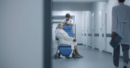 Photo for Male doctor stands with female patient in wheelchair in clinic corridor. Medic talks to elderly woman before surgery near operating room. Medical staff, patients in hospital or medical center hallway. - Royalty Free Image