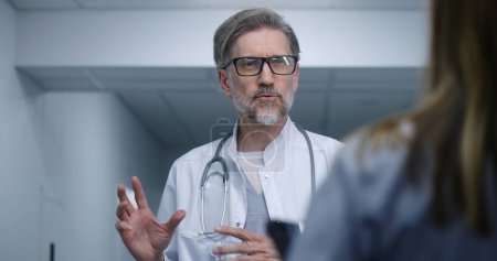 Photo for Mature doctor stands in modern clinic corridor, talks with female colleague or patient. Healthcare specialist discusses treatment or diagnostic results. Medical staff and patients in hospital hallway. - Royalty Free Image