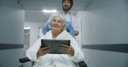 Foto de Mujer mayor observa los resultados de las pruebas médicas en la tableta digital y lo discute con un médico joven. Médico profesional lleva a viejo paciente en silla de ruedas por el pasillo del centro médico. - Imagen libre de derechos