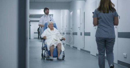 Foto de Enfermera masculina empuja silla de ruedas con paciente femenino en corredor clínico. Mujer médico viene a colega y paciente de edad avanzada, habla, utiliza tableta digital. Personal médico y pacientes en el pasillo del hospital. - Imagen libre de derechos