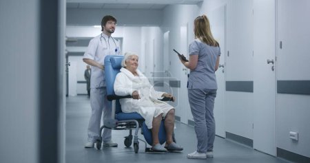 Krankenschwester schiebt Rollstuhl mit Patientin im Klinikflur. Ärztin kommt zu Kollegin und älterer Patientin, spricht, nutzt digitales Tablet. Medizinisches Personal und Patienten im Krankenhausflur.