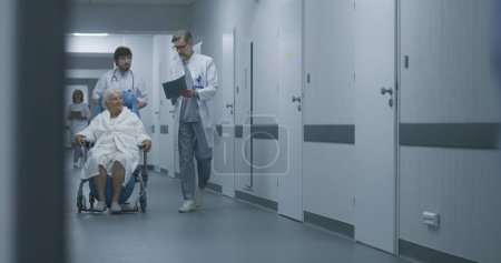Photo for Back view of doctors pushing transfer wheelchair with patient walking along corridor. Medics talk, take person on procedures, therapy or to hospital room. Medical staff and patients in clinic hallway. - Royalty Free Image