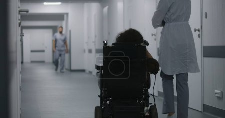 Foto de La doctora camina por el pasillo de la clínica, consulta a una mujer con discapacidad física. El médico habla sobre los procedimientos médicos con el paciente en silla de ruedas eléctrica. Corredor de las instalaciones médicas modernas. - Imagen libre de derechos