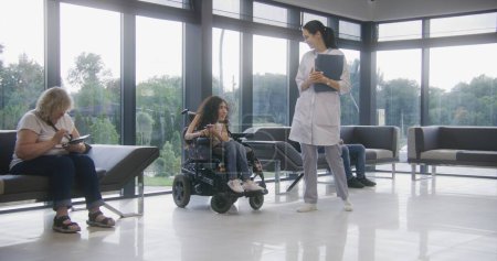 Téléchargez les photos : Une femme en fauteuil roulant électrique attend la consultation d'un médecin dans la salle d'attente de la clinique. Infirmière invite le patient handicapé sur les procédures. Personnel médical et personnes dans le hall des installations médicales modernes. - en image libre de droit