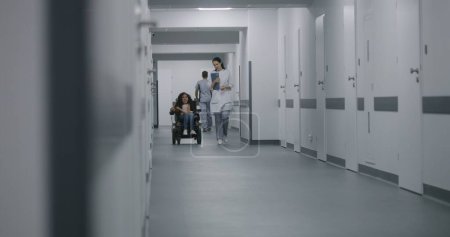Foto de La doctora camina por el pasillo de la clínica, consulta a la paciente con discapacidad física. Médico habla de chequeo médico con mujer en silla de ruedas motorizada. Pasillo de las instalaciones médicas modernas. - Imagen libre de derechos