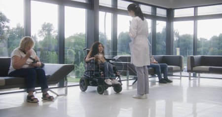 Photo for Woman in electric wheelchair waits for consultation with doctor in clinic waiting area. Nurse invites patient with disability on procedures. Medical staff and people in modern medical facility lobby. - Royalty Free Image