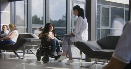Téléchargez les photos : Femme handicapée en fauteuil roulant motorisé parle au médecin dans le hall de l'hôpital ou de la clinique moderne. Médecin professionnel assis sur le canapé, consulte une patiente atteinte d'atrophie musculaire de la colonne vertébrale. Dolly shot. - en image libre de droit
