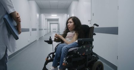 Téléchargez les photos : Couloir de l'hôpital : Femme handicapée SMA en fauteuil roulant parle à un médecin professionnel de contrôle médical. Femme médecin consulte le patient dans le couloir lumineux de la clinique moderne ou d'un établissement médical. - en image libre de droit