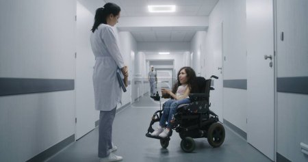 Téléchargez les photos : Couloir de l'hôpital : Femme handicapée SMA en fauteuil roulant parle à un médecin professionnel de contrôle médical. Femme médecin consulte le patient dans le couloir lumineux de la clinique moderne ou d'un établissement médical. - en image libre de droit