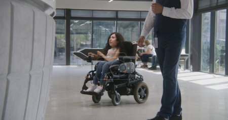 Téléchargez les photos : Femme handicapée SMA monte à l'entrée de la clinique en fauteuil roulant motorisé à la réception et parle avec l'administrateur. Un travailleur médical rencontre une patiente. Lobby de l'hôpital ou de l'établissement médical moderne. - en image libre de droit