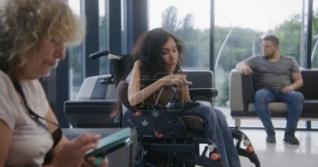 Foto de Mujer con discapacidad en silla de ruedas motorizada espera cita con el médico en el vestíbulo de la clínica, utiliza el teléfono móvil. Diversas personas se sientan en sofás en el hospital o en la moderna sala de espera de las instalaciones médicas. - Imagen libre de derechos