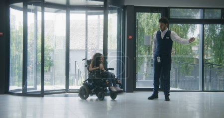 Photo for Woman with SMA disability rides in clinic entrance on motorized wheelchair through revolving doors. Male administrator meets and greets female patient. Hospital or modern medical facility lobby area. - Royalty Free Image