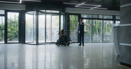 Femme handicapée SMA monte à l'entrée de la clinique sur fauteuil roulant motorisé par des portes tournantes. Homme administrateur rencontre et accueille la patiente. Hall d'entrée hôpital ou établissement médical moderne.