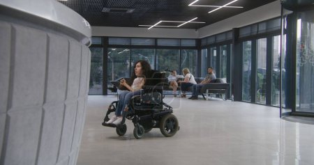 Téléchargez les photos : Femme handicapée SMA monte à l'entrée de la clinique en fauteuil roulant motorisé à la réception et parle avec l'administrateur. Un travailleur médical rencontre une patiente. Lobby de l'hôpital ou de l'établissement médical moderne. - en image libre de droit