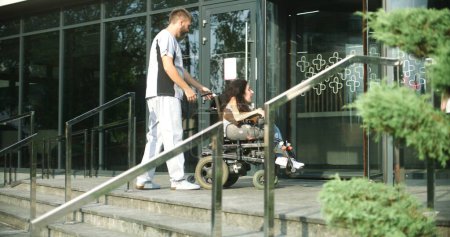 Foto de Mujer con discapacidad física entra en la entrada de la clínica en silla de ruedas motorizada. Trabajador médico, enfermero se reúne y ayuda a la paciente a entrar. Hospital o centro médico moderno fuera. - Imagen libre de derechos