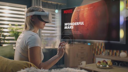 Photo for Woman in VR headset chooses movie or TV show to watch at home. 3D futuristic hologram shows interface of streaming service application and widgets in user menu. VFX animation. Concept of lifestyle. - Royalty Free Image