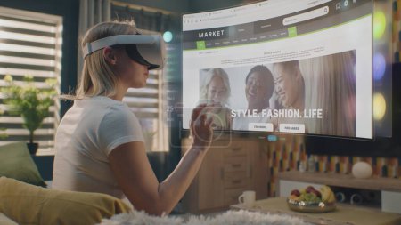 Téléchargez les photos : Femme en VR casque choisit des vêtements dans la boutique en ligne refroidissant à la maison. L'hologramme futuriste 3D montre les widgets dans le menu utilisateur et l'interface du site Web du magasin de vêtements. Animation VFX. Concept d'achat en ligne. - en image libre de droit