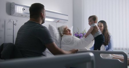 Téléchargez les photos : Une patiente âgée se couche dans un hôpital lumineux, joue avec son petit-fils. Les membres aimants de la famille aident les femmes âgées à se rétablir après une chirurgie réussie. Installation médicale moderne. - en image libre de droit
