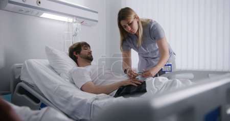 Photo for Female nurse comes to resting in bed patient, puts on heart rate monitor on finger and corrects medical drip. Man fully recovering after successful surgery. Medical personnel work in hospital ward. - Royalty Free Image