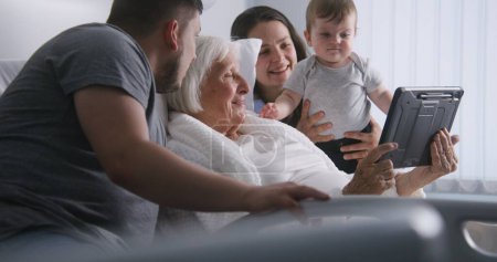 Photo for Hospital room. Old woman lying in bed spends time with relatives. Loving family use tablet computer, support elderly grandmother recovering after successful surgery. Modern medical facility or clinic. - Royalty Free Image