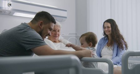 Photo for Elderly woman lying and resting in bed in bright hospital ward, plays with grandson. Loving family members support grandmother recovering after successful surgery. Modern medical facility or clinic. - Royalty Free Image