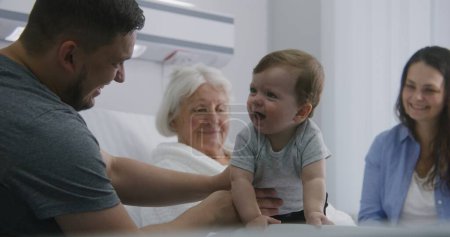 Photo for Old woman resting in bed in bright hospital room, plays with little grandson. Loving family supports grandmother recovering after successful surgery. Modern medical facility or clinic. Slow motion. - Royalty Free Image