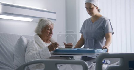 Téléchargez les photos : Infirmière ouvre une grande boîte à lunch avec repas à une patiente âgée. Femme âgée se reposant au lit dans une chambre d'hôpital lumineuse, se rétablissant après une chirurgie réussie, mangeant de la nourriture. Installation médicale ou clinique moderne. - en image libre de droit