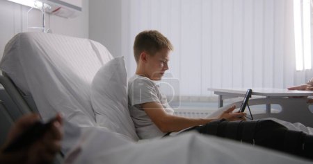 Photo for Young boy with bandaged leg lies on bed, talks with dad using tablet. Kid remotely shows hospital conditions to father. Patient fully recovering after successful surgery. Modern and bright clinic. - Royalty Free Image