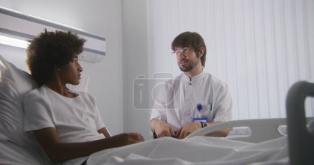 Téléchargez les photos : Adolescent afro-américain et son père parlent avec un médecin caucasien de la santé dans la chambre d'hôpital. Médecin adulte avec tablette numérique consulte garçon dans la salle d'hôpital. Personnel médical au travail en clinique. - en image libre de droit